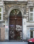 Wejście do budynku, w którym mieści się dzienny dom pomocy. Drewniana brama zwieńczona szklanym łukiem. Przed wejściem czerwony samochód